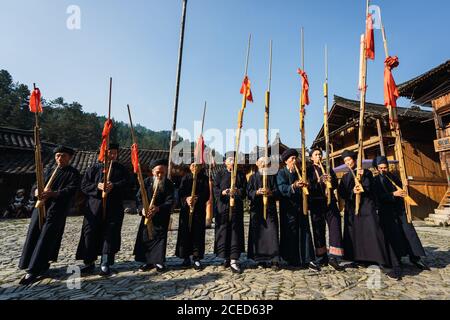 GUINZHOU, CHINA - 14. JUNI 2018: Gruppe von männlichen Musikern der ethnischen Minderheit Miao in dunklen langen Kostümen und Kappen, die traditionelle Instrumente spielen, stehen am sonnigen Tag auf dem Dorfplatz in der chinesischen Provinz Guizhou Stockfoto