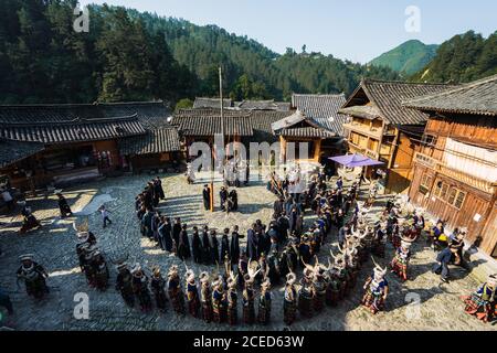 GUINZHOU, CHINA - 14. JUNI 2018: Luftaufnahme einer großen Menschenmenge von Miao ethnischen Gruppe Menschen in traditionellen Kostümen spielen Musikinstrumente und tanzen stehend im Halbkreis auf Dorfstraße mit alten Holzdielen Häuser in der Provinz Guizhou in China Stockfoto