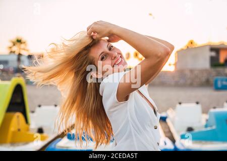 Seitenansicht der fröhlichen jungen Blondine?weiblich in weißen Sommerhemd Stehen mit dem Kopf zurück berühren Haare und Blick auf die Kamera Fröhlich lächelnd auf verschwommenem Hintergrund beleuchtet Strand Sand Und Boote bei Sonnenuntergang Stockfoto