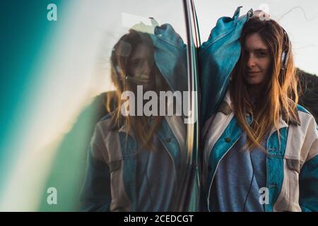 Ziemlich lässig gekleidete Frau, die sich auf das Auto lehnt und Haare in der Natur anfasst. Stockfoto