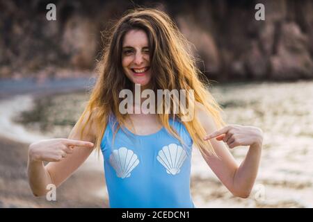 Winkende und lächelnde Frau zeigt auf Muscheldruck auf Badeanzug am Strand. Stockfoto