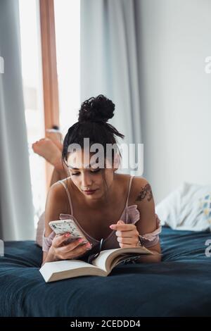 Hübsche Brünette entspannend zu Hause liegend auf dem Bett mit Buch Geöffnet und mit Smartphone in der Freizeit Stockfoto