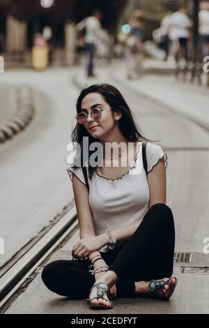 Junge Hipster Mädchen in Sandalen und Sonnenbrillen sitzen mit Rucksack Auf asphaltierter Straßenbahnstraße in der Stadt Stockfoto