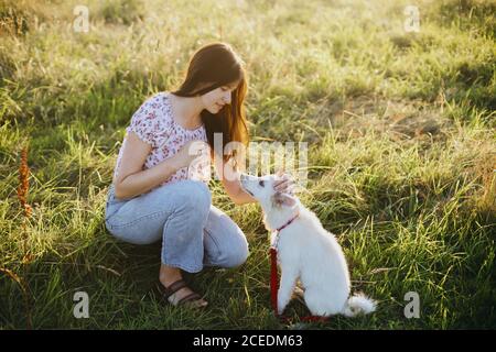 Frau Ausbildung niedlichen weißen Welpen zu benehmen und streicheln ihn in Sommerwiese in warmen Sonnenuntergang Licht. Liebenswert flauschig Welpen Blick auf Mädchen Besitzer. Loyal Stockfoto