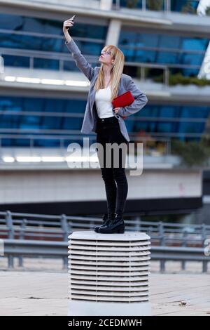 Attraktive Frau in grauer Jacke und schwarzer Jeans macht Foto am Telefon mit rotem Lippenstift Stockfoto