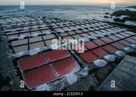 Malerischer Blick auf kleine rechteckige Kristallisationsteiche mit Meerwasser Und produzierte Salz in Pfählen rund um an der felsigen Küste Bei Sonnenuntergang in Spanien Stockfoto