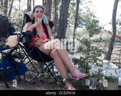 Picknick oder Ruhe im Wald. Attraktive Frau lacht in einem Wanderstuhl sitzend hält sie ein Glas, genießt sie eine Ruhe in der Natur Stockfoto