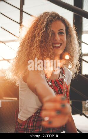 Wunderbare junge lockige Frau lächelt der Kamera zu, während sie einen verschwommenen glühenden Stock zeigt, der bei Sonnenschein auf der Treppe sitzt Stockfoto