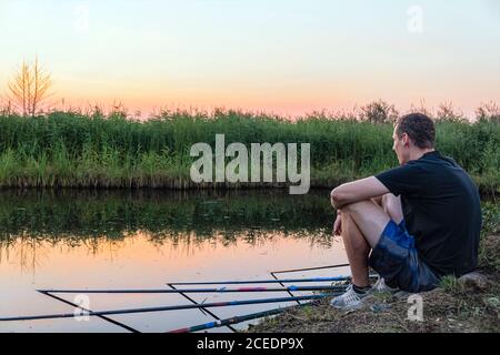 Ruhiger entspannter Mann fängt Angeln auf Angelruten sitzen auf Das Ufer des Sees bei Sonnenuntergang Stockfoto