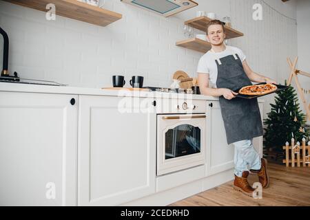Hausgemachte italienische Pizza, schöner junger Koch in dunkler Schürze hält Backblech in heller Küche. Kochen Video Blog Konzept Stockfoto