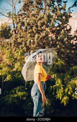 Attraktive junge Frau im stilvollen Casual Outfit fröhlich lächelnd und Blick auf die Kamera, während Sie den nassen Regenschirm halten und darauf stehen Straße in der Nähe von Büschen Stockfoto