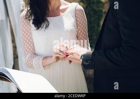 Crop-Ansicht der jungen Braut in Hochzeitskleid und Bräutigam In schwarzem Kostüm tauschen Ringe bei Zeremonie im Garten Stockfoto