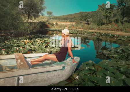 Unbeschwerte junge Frau, die entspannt auf einem Boot an einem Teich mit blühenden Seerosen, Lotusblumen schwimmt. Urlaubsreise, natürlicher Hintergrund am See mit Stockfoto