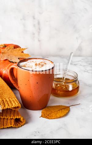 Kürbisse würzen Latte oder Kaffee mit cremigem Schaum in orangefarbener Tasse mit Kürbis, Blättern, Glas Honig und weißem gemütlichen Pullover auf weißem Marmor Hintergrund Stockfoto