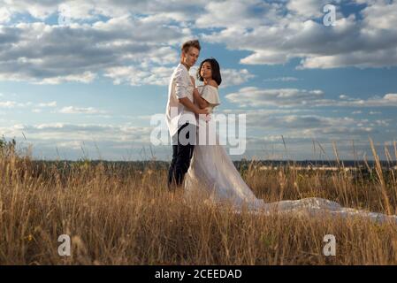 Junges Paar Braut und Bräutigam im Hochzeitskleid, weißes Kleid, auf dem Feld, vor dem Hintergrund des wolkigen blauen Himmels. Hochzeitsaufnahmen Stockfoto