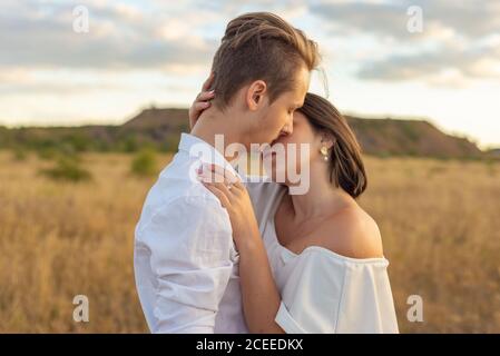 Kuss von einem Mädchen und einem Kerl. Nahaufnahme Porträt. Junges Paar von Kerl und Mädchen in weißen Kleidern, auf dem Feld. Vor dem Hintergrund eines bewölkten Himmels Stockfoto