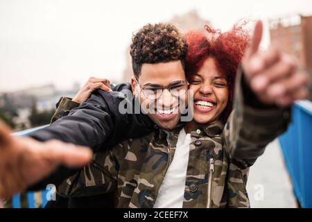 Aufgeregt lachendes afroamerikanisches Paar auf der Straße umarmt und ausstreckt Hände Bei der Kamera in Spaß Stockfoto