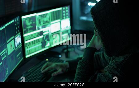 Gefährlicher Hacker mit Kapuze an seinem Versteck, der eine dunkle Atmosphäre, mehrere Displays hat Stockfoto