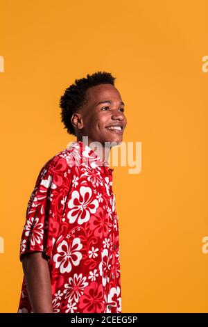 Seitenansicht eines lächelnden jungen afroamerikanischen Mannes, der einen farbenfrohen Strand trägt shirt und Blick weg auf orangenen Hintergrund Stockfoto