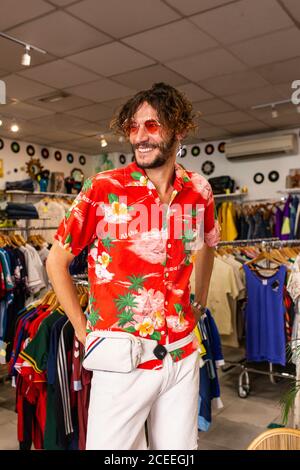 Schöner Mann in hellen stilvollen Outfit lächelnd und wegschauen Während Sie in einem kleinen Kleiderladen stehen Stockfoto