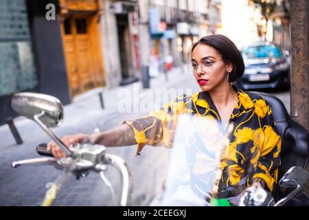 Schöne junge Frau im trendigen Outfit sitzt auf Motorrad auf Verschwommener Hintergrund der Stadtstraße Stockfoto