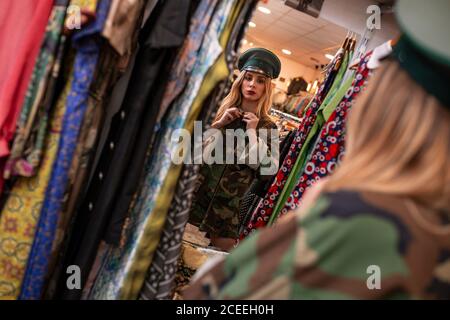 Schöne junge Frau, die in der Nähe des Spiegels steht und beim Militär versucht Kleidung, während Sie Zeit in einem kleinen Laden verbringen Stockfoto