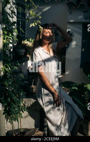 Seitenansicht einer hübschen Frau in weißem Kleid, die im Schatten in der Nähe des Landhauses steht und in der Ferne blickt Stockfoto