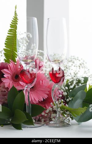 Hochzeitsgläser mit roten Herzen sind auf dem Fenster, Eheringe im Inneren und ein Strauß rosa Blumen Stockfoto