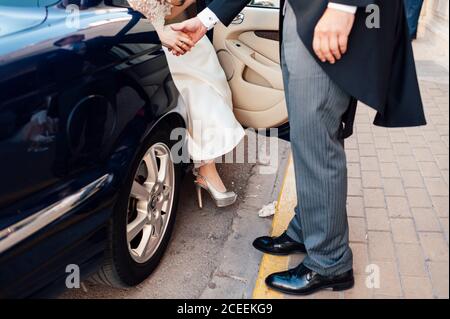 Crop Mann im Anzug hilft Braut zu Fuß aus dem schwarzen Auto. Stockfoto