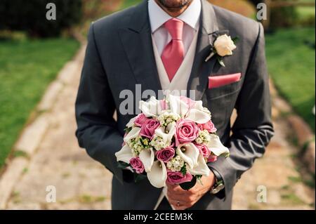 Crop schöner Mann in grauem Anzug stehen mit Bündel von rosa und weißen Blumen. Stockfoto