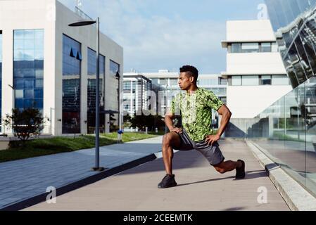 Schöner muskulöser ethnischer Mann, der sich auf der Straße der modernen Stadt ausdehnt und die Beine erwärmt. Stockfoto