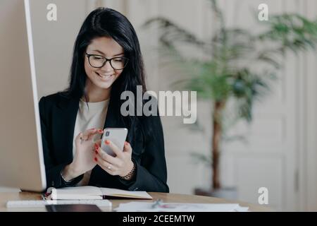 Schöne lächelnde Freiberuflerin arbeitet aus der Ferne, konzentriert in Smartphone, sendet Feedback auf empfangene Nachricht, sitzt im Coworking Space, gekleidet f Stockfoto