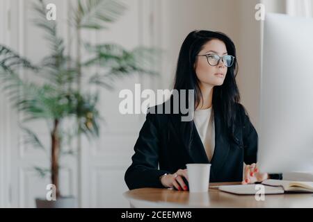 Konzentrierte Unternehmerin trägt elegantes schwarzes Kostüm, arbeitet am Computer, sitzt am Tisch in gemütlichen Arbeitsplatz trägt transparente Gläser Browses in Stockfoto