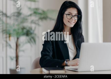 Lächelnd gut gekleidete junge Frau Sekretärin mit dunklen Haaren, arbeitet auf Laptop-Computer am Desktop, macht Projekt oder Forschung Arbeit. Vertriebsleiter in Modder Stockfoto