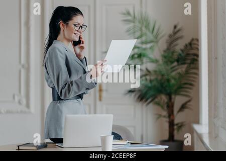 Innenaufnahme der glücklich lächelnden Frau Manager prüft Informationen aus Papieren trägt graue formale Kostüm, optische Brille auf Forschungsarbeit konzentriert hat Stockfoto