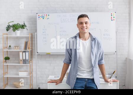 Online englischkurse zu Hause. Lächelnder Kerl steht in der Nähe von Tisch und Brett im Inneren und lehrt Studenten aus der Ferne Stockfoto