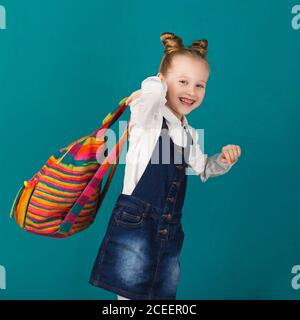 Lustige lächelnde kleine Mädchen mit großen Rucksack springen und Spaß an der blauen Wand. Blick auf die Kamera. Schulkonzept. Zurück zur Schule Stockfoto