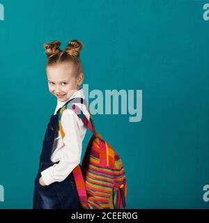 Schönes lächelndes kleines Mädchen mit großem Strickrucksack, der gegen die blaue Wand steht. Blick auf die Kamera. Schulkonzept. Zurück zur Schule Stockfoto