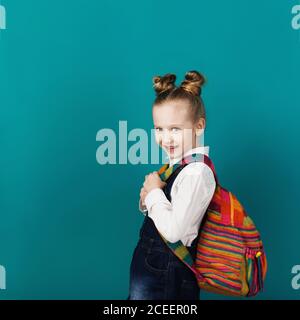 Schönes lächelndes kleines Mädchen mit großem Rucksack, der gegen die blaue Wand steht. Blick auf die Kamera. Schulkonzept. Zurück zur Schule Stockfoto