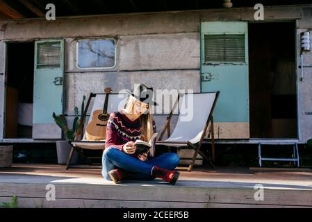 Charmante junge Frau in stilvollem Outfit lesen Interessantes Buch, während auf der Terrasse in der Nähe von gealterten Wohnmobil sitzen Stockfoto
