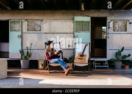 Charmante junge Frau in stilvollem Outfit lesen Interessantes Buch, während auf der Terrasse in der Nähe von gealterten Wohnmobil sitzen Stockfoto