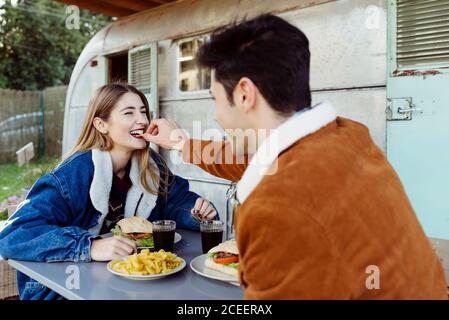 Seitenansicht eines jungen Mannes und einer Frau, die lächelten und einander anschauten, während sie in der Nähe eines schäbigen Wohnwagens ein köstliches romantisches Abendessen aßen Stockfoto