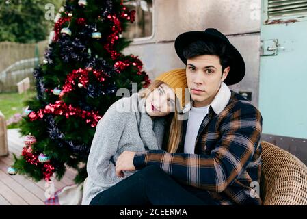 Fröhlicher junger Mann und Frau umarmen sich gegenseitig, während sie in einem Sessel neben einem schönen Weihnachtsbaum vor einem alten Lieferwagen sitzen Stockfoto