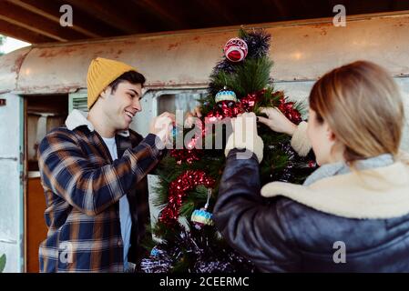 Junger Mann und Frau in stilvollen Kleidern schmücken schönen Weihnachtsbaum in der Nähe von gealterten Wohnwagen auf dem Land Stockfoto