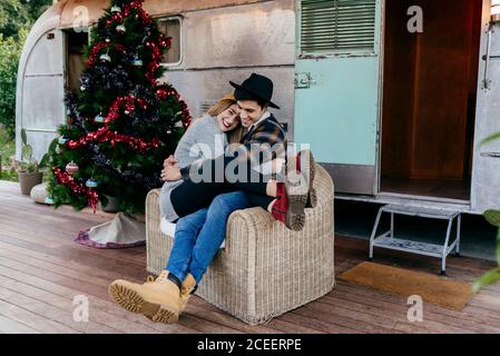 Fröhlicher junger Mann und Frau umarmen sich gegenseitig, während sie in einem Sessel neben einem schönen Weihnachtsbaum vor einem alten Lieferwagen sitzen Stockfoto