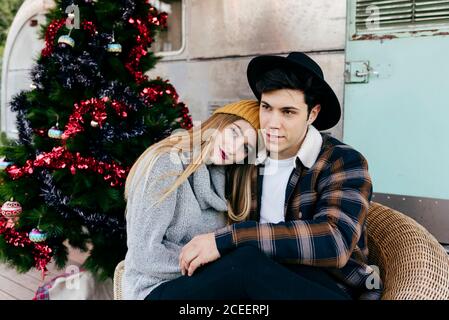 Fröhlicher junger Mann und Frau umarmen sich gegenseitig, während sie in einem Sessel neben einem schönen Weihnachtsbaum vor einem alten Lieferwagen sitzen Stockfoto