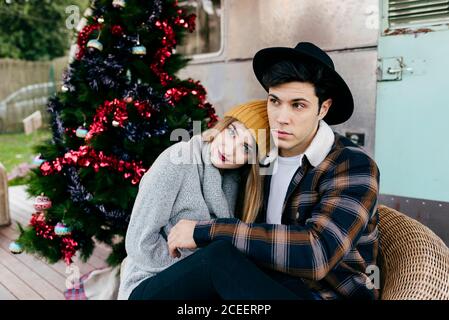Fröhlicher junger Mann und Frau umarmen sich gegenseitig, während sie in einem Sessel neben einem schönen Weihnachtsbaum vor einem alten Lieferwagen sitzen Stockfoto
