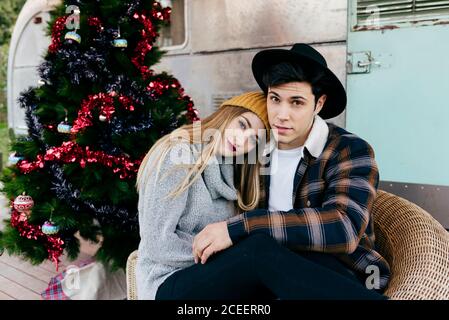 Fröhlicher junger Mann und Frau umarmen sich gegenseitig, während sie in einem Sessel neben einem schönen Weihnachtsbaum vor einem alten Lieferwagen sitzen Stockfoto