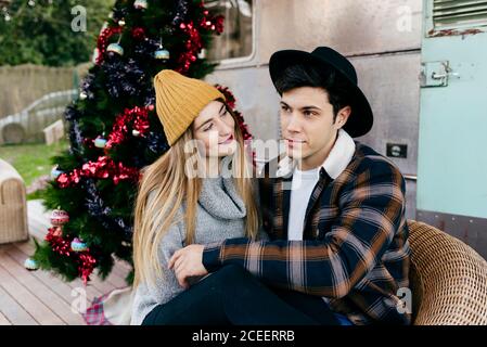 Fröhlicher junger Mann und Frau umarmen sich gegenseitig, während sie in einem Sessel neben einem schönen Weihnachtsbaum vor einem alten Lieferwagen sitzen Stockfoto