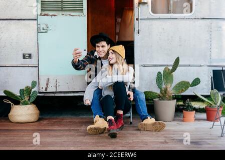 Hübscher junger Mann und schöne Frau, die lächeln und für Selfie posieren, während sie in der Nähe eines verwitterten Wohnwagens sitzen Stockfoto